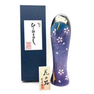 May include: A traditional Japanese wooden doll, known as a kokeshi, with a purple and blue kimono decorated with white cherry blossoms. The doll is standing on a white surface and has a small wooden tag with Japanese text that reads "Flower of the Night".