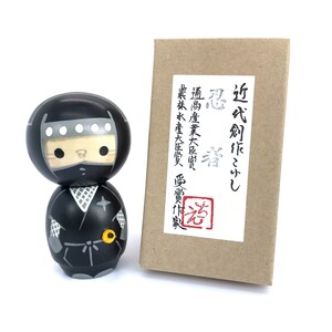 May include: A black and white kokeshi doll depicting a ninja. The doll is wearing a black mask and a black and white ninja outfit. The doll is standing on a white surface. The doll is in front of a brown box with Japanese text that reads "近代創作こけし 忍者", which translates to "Modern Creation Kokeshi Ninja".