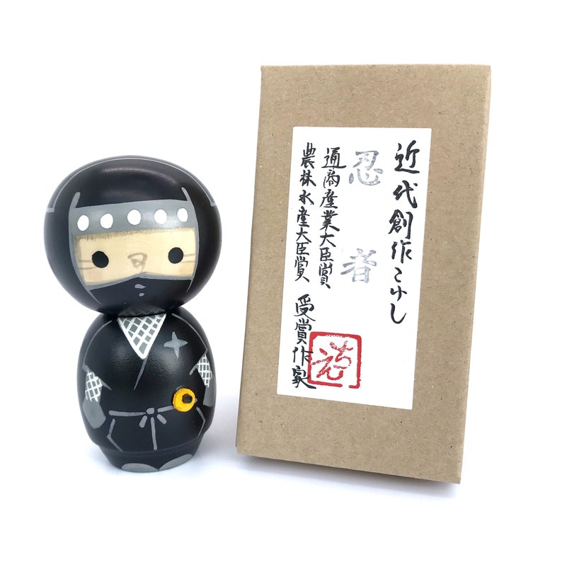May include: A black and white kokeshi doll depicting a ninja. The doll is wearing a black mask and a black and white ninja outfit. The doll is standing on a white surface. The doll is in front of a brown box with Japanese text that reads "近代創作こけし 忍者", which translates to "Modern Creation Kokeshi Ninja".