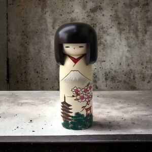 May include: A traditional Japanese kokeshi doll with black hair and a white kimono. The doll is painted with a scene of Mount Fuji, cherry blossoms, a pagoda, and a torii gate.