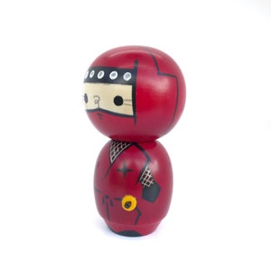 May include: A red wooden doll with a black ninja outfit and a white face. The doll has a black eye mask and a white star on its chest.