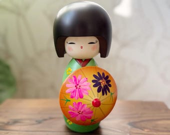 May include: A wooden Kokeshi doll with a black helmet-shaped head and a green kimono. The doll holds a yellow parasol decorated with pink and purple flowers. The doll is standing on a wooden surface.
