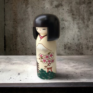 May include: A traditional Japanese kokeshi doll with black hair, a white face, and a painted design of a mountain, cherry blossoms, and a red torii gate. The doll is made of wood and has a smooth, polished finish.