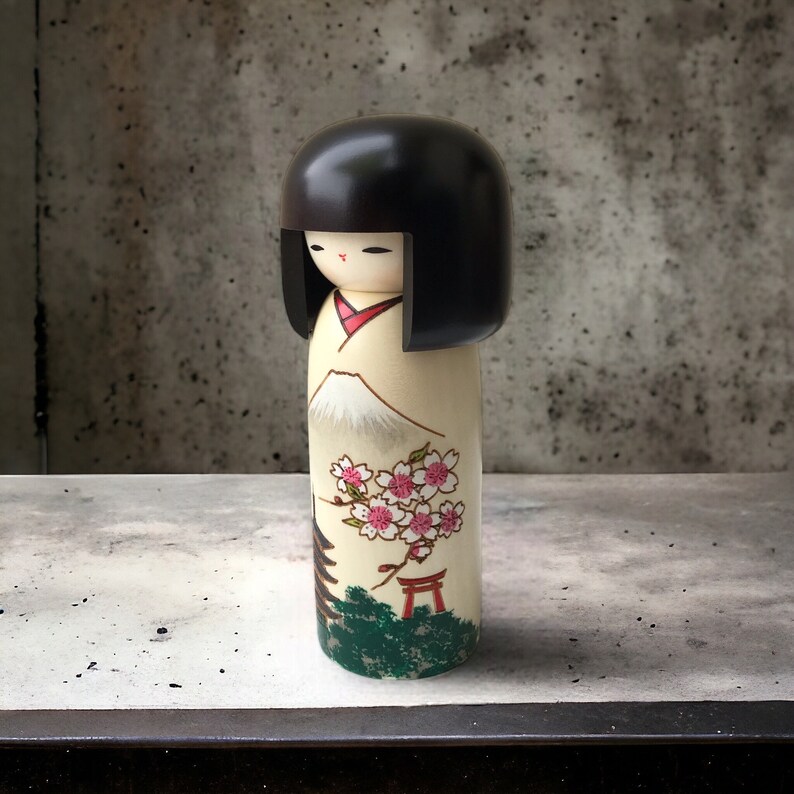 May include: A traditional Japanese kokeshi doll with black hair, a white face, and a painted design of a mountain, cherry blossoms, and a red torii gate. The doll is made of wood and has a smooth, polished finish.
