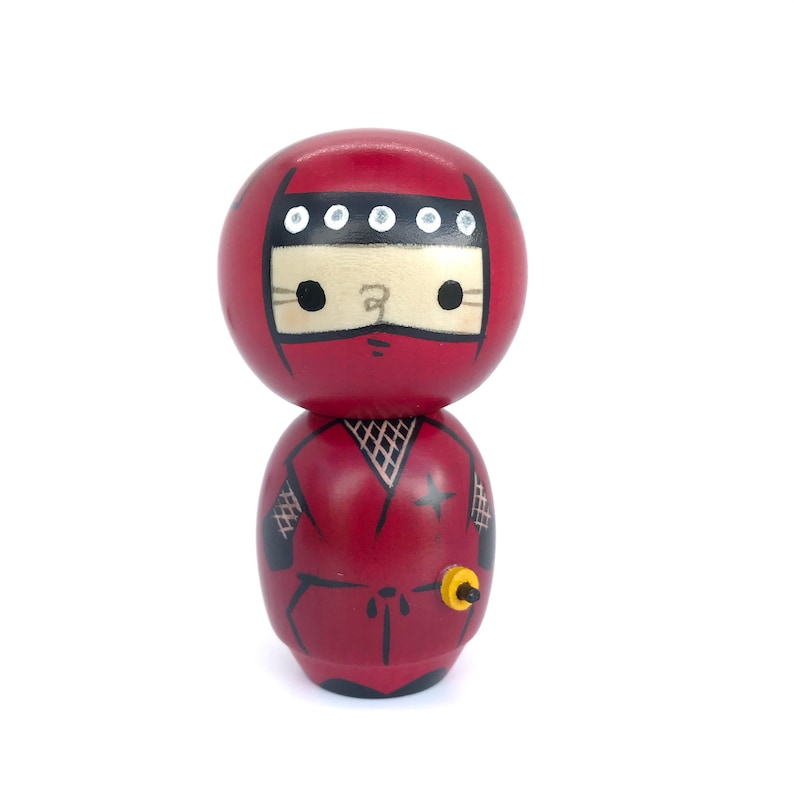 May include: A red wooden doll with a black ninja outfit and a black headband with white dots. The doll has a black star on its chest and a yellow sword.