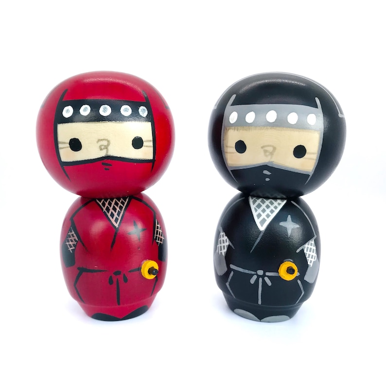 May include: Two wooden figurines of ninjas, one in red and one in black, with white eyes and a black headband. Both figurines have a black star on their chest and a yellow star on their belt.