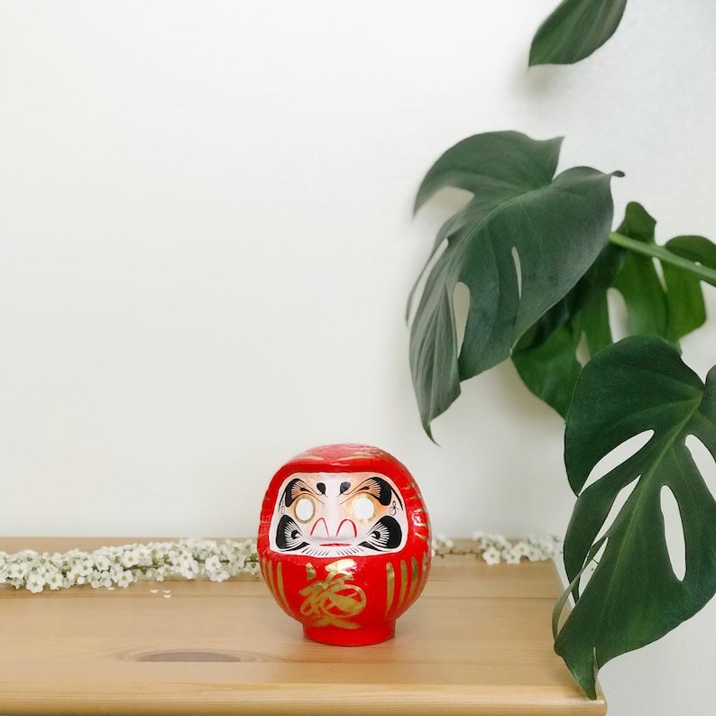 May include: A red and gold Japanese lucky charm, known as a Daruma doll, with a white face and black eyes. The doll is sitting on a wooden surface with a green plant in the background.