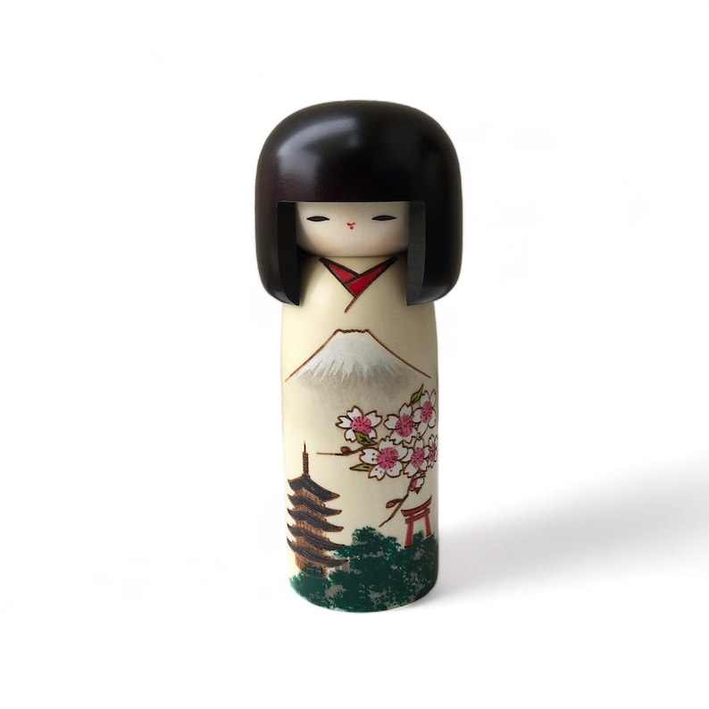May include: A traditional Japanese kokeshi doll with black hair, a red and white kimono, and a painted scene of Mount Fuji, cherry blossoms, a pagoda, and a torii gate.