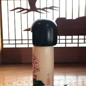 May include: A traditional Japanese kokeshi doll with a black hairpiece and a white body. The doll is decorated with pink cherry blossoms and a red torii gate. The doll is standing on a wooden surface.