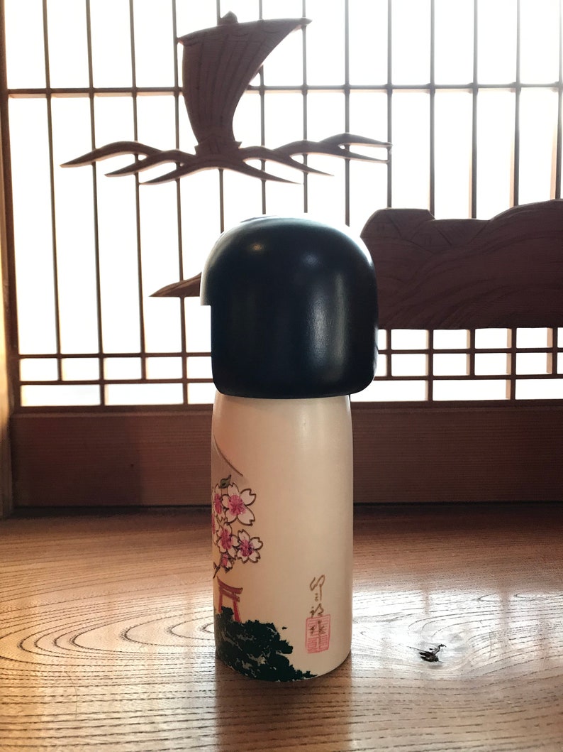 May include: A traditional Japanese kokeshi doll with a black hairpiece and a white body. The doll is decorated with pink cherry blossoms and a red torii gate. The doll is standing on a wooden surface.