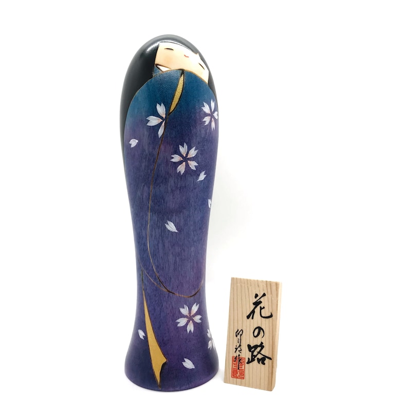 May include: A wooden Japanese kokeshi doll with a purple and blue painted kimono with white cherry blossoms. The doll is standing on a white background. The doll has a small wooden tag with Japanese characters that read "Flower of the Night".