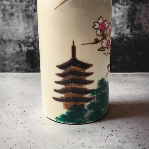 May include: A white ceramic bottle with a Japanese pagoda and cherry blossoms design. The pagoda is black and gold, and the cherry blossoms are pink and white.