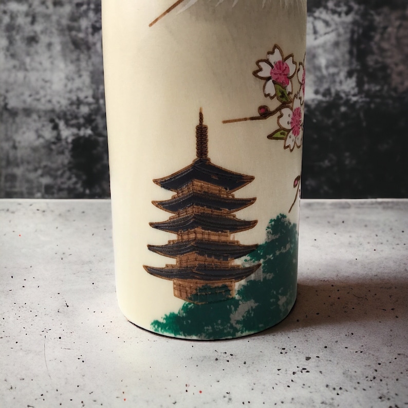 May include: A white ceramic bottle with a Japanese pagoda and cherry blossoms design. The pagoda is black and gold, and the cherry blossoms are pink and white.