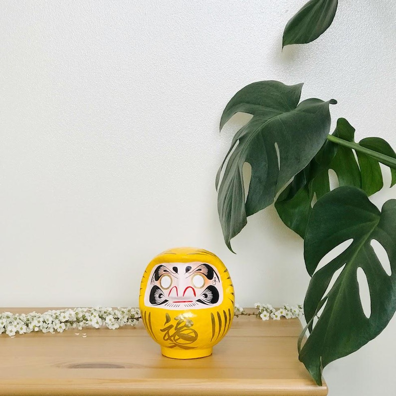 May include: A yellow Japanese lucky charm, known as a Daruma doll, with a painted face and gold accents. The doll is sitting on a wooden surface with white flowers in the background.