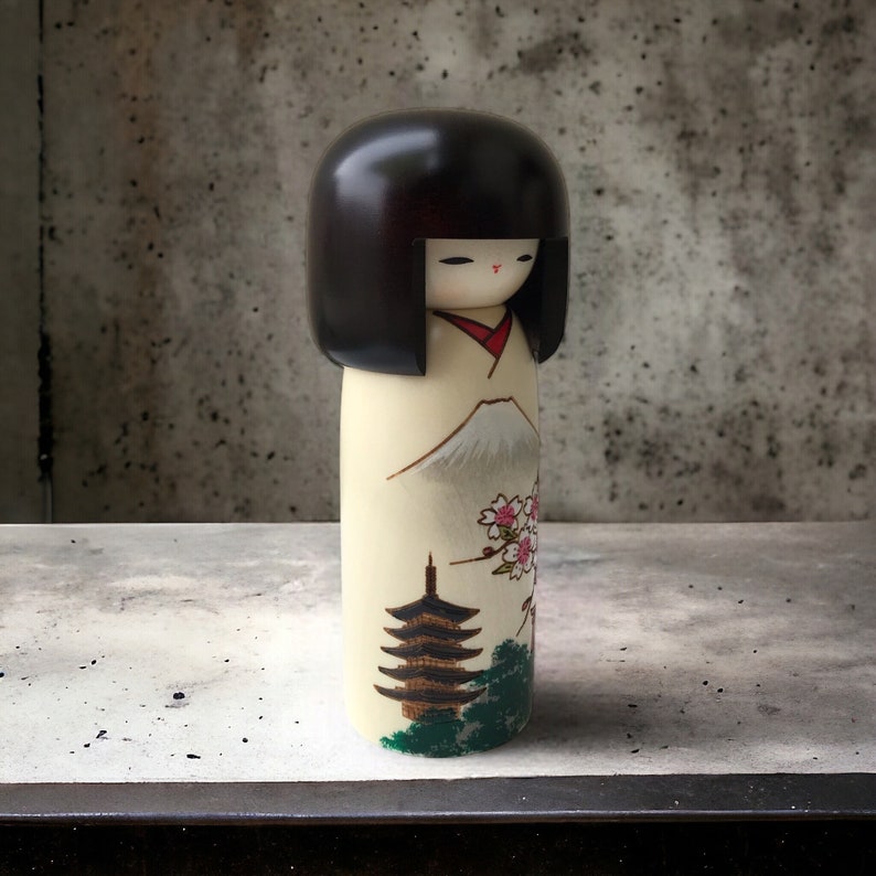 May include: A traditional Japanese kokeshi doll with black hair, a white kimono, and a painted scene of Mount Fuji, cherry blossoms, and a pagoda.