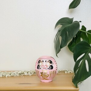 May include: Pink and gold Japanese lucky charm, or "daruma" doll, with a black and white face and the kanji character for "luck" written in gold on the front. The doll is sitting on a wooden table with a green plant in the background.