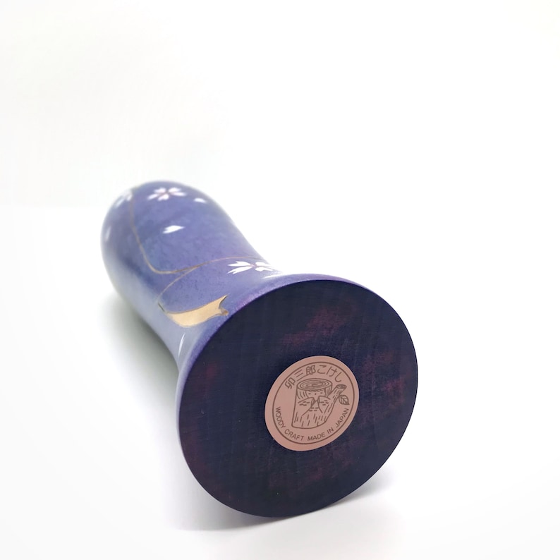 May include: A purple wooden vase with a white and gold floral design. The vase has a round base and a label on the bottom that reads "Woody Craft Made in Japan".