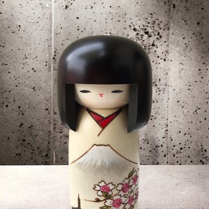 May include: A traditional Japanese kokeshi doll with black hair, a white and red kimono, and a painted design of Mount Fuji and cherry blossoms.