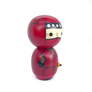 May include: A red wooden doll with a black ninja outfit and a black headband with white dots. The doll is holding a small yellow object in its right hand.