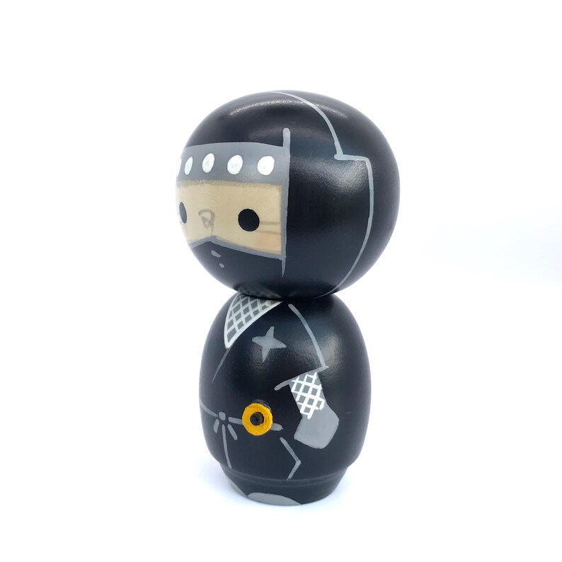 May include: A black and gray wooden doll with a ninja costume. The doll has a black hood with white accents and a black mask. The doll is wearing a black and gray ninja outfit with a star on the chest.