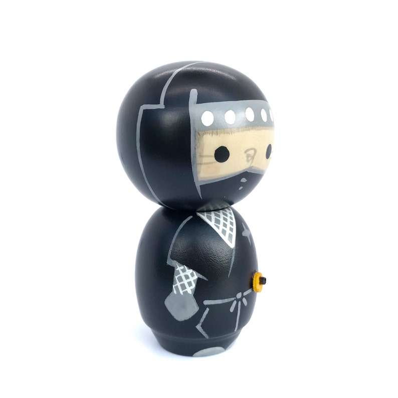 May include: A black wooden doll with a black ninja outfit and a white mask. The doll has a yellow detail on the side of the outfit.