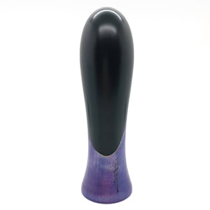 May include: A black and purple wooden bottle stopper with a rounded top and a flared base. The base is painted with a purple and blue gradient and features a black signature.