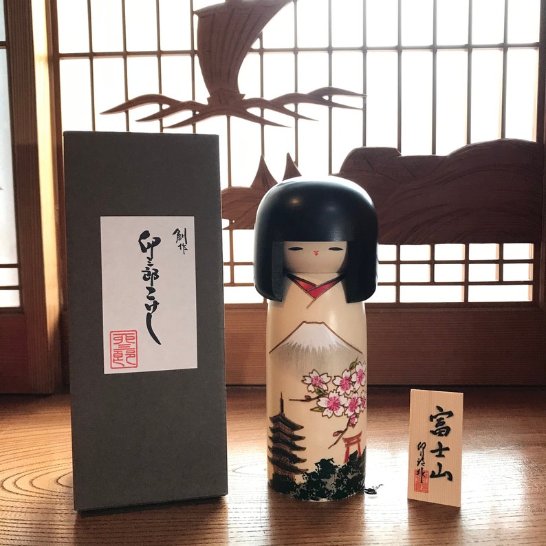 May include: A traditional Japanese kokeshi doll with a black hairpiece and a painted face. The doll is wearing a white kimono with a red sash and a mountain scene with cherry blossoms and a pagoda. The doll is in a box with Japanese writing on it.