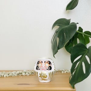 May include: White ceramic Japanese lucky charm with a gold and black painted design. The charm has a round body and a flat base. The charm is sitting on a wooden surface. The charm has the Japanese character for "good fortune" written on it.