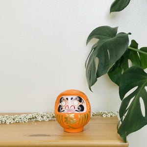 May include: Orange and gold Japanese lucky charm, also known as a Daruma doll, with a white face and black eyes. The doll is sitting on a wooden surface with white flowers in the background.
