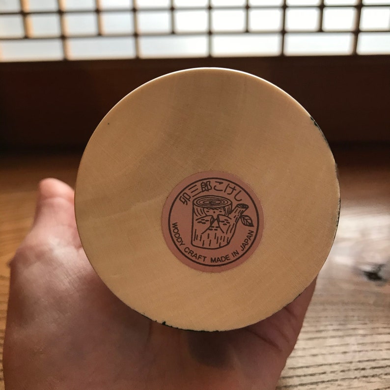 May include: A close-up of the bottom of a wooden craft item. The item is light brown and has a round base. A brown label with the text "Woody Craft Made in Japan" and a stylized tree stump design is attached to the base.