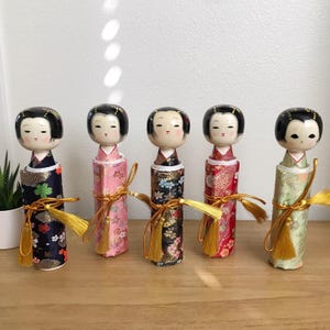 May include: Five traditional Japanese dolls, known as kokeshi, are arranged in a row. Each doll is made of wood and painted with intricate designs. The dolls are dressed in colorful kimonos and have black hair. The dolls are tied with gold ribbons and have gold tassels.
