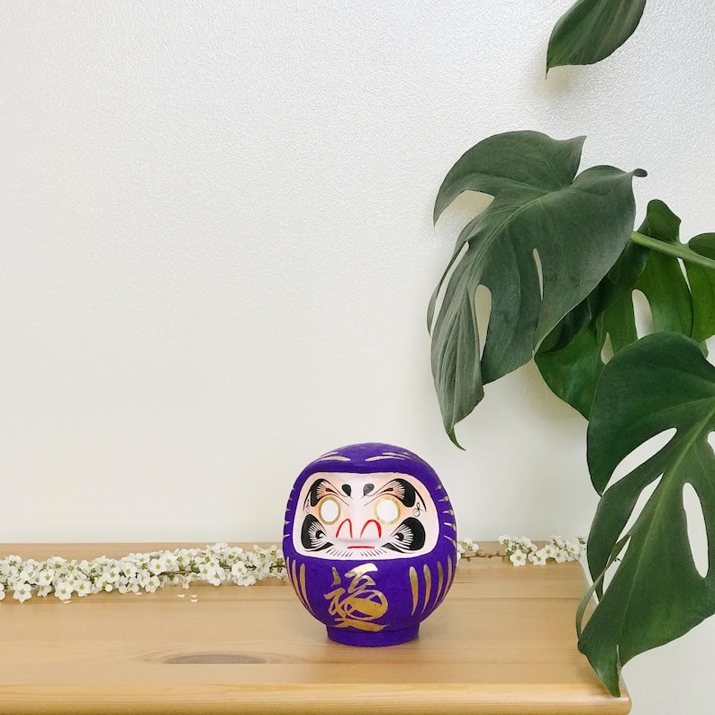 May include: A purple and gold Japanese lucky charm, a daruma doll, with a white face and gold accents. The doll is sitting on a wooden surface with a white background.