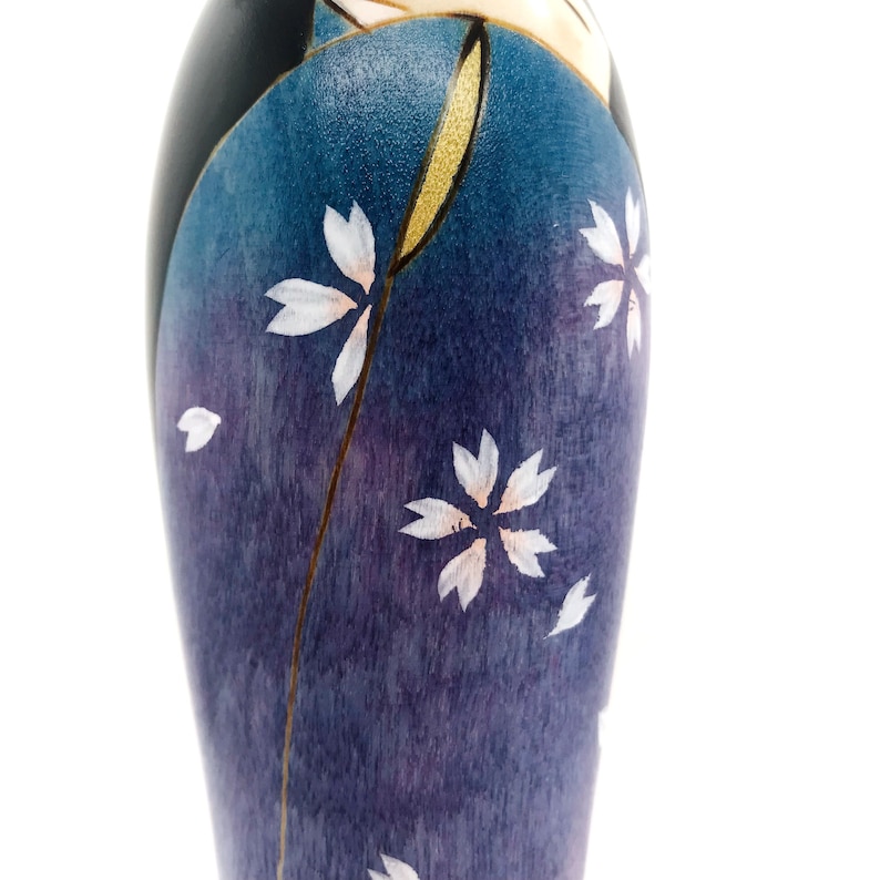 May include: A wooden doll with a blue and purple patterned kimono. The kimono is decorated with white flowers and gold accents.