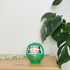 May include: A green and gold Japanese lucky charm, known as a Daruma doll, with a white face and black eyes. The doll is sitting on a wooden surface.