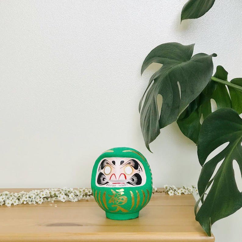 May include: A green and gold Japanese lucky charm, known as a Daruma doll, with a white face and black eyes. The doll is sitting on a wooden surface.