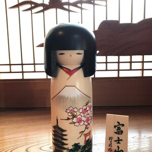 May include: A traditional Japanese kokeshi doll with a black hair, red lips, and a white body with a painted scene of Mount Fuji, cherry blossoms, a pagoda, and a torii gate. The doll is standing on a wooden surface with a small wooden sign that reads "富士山".