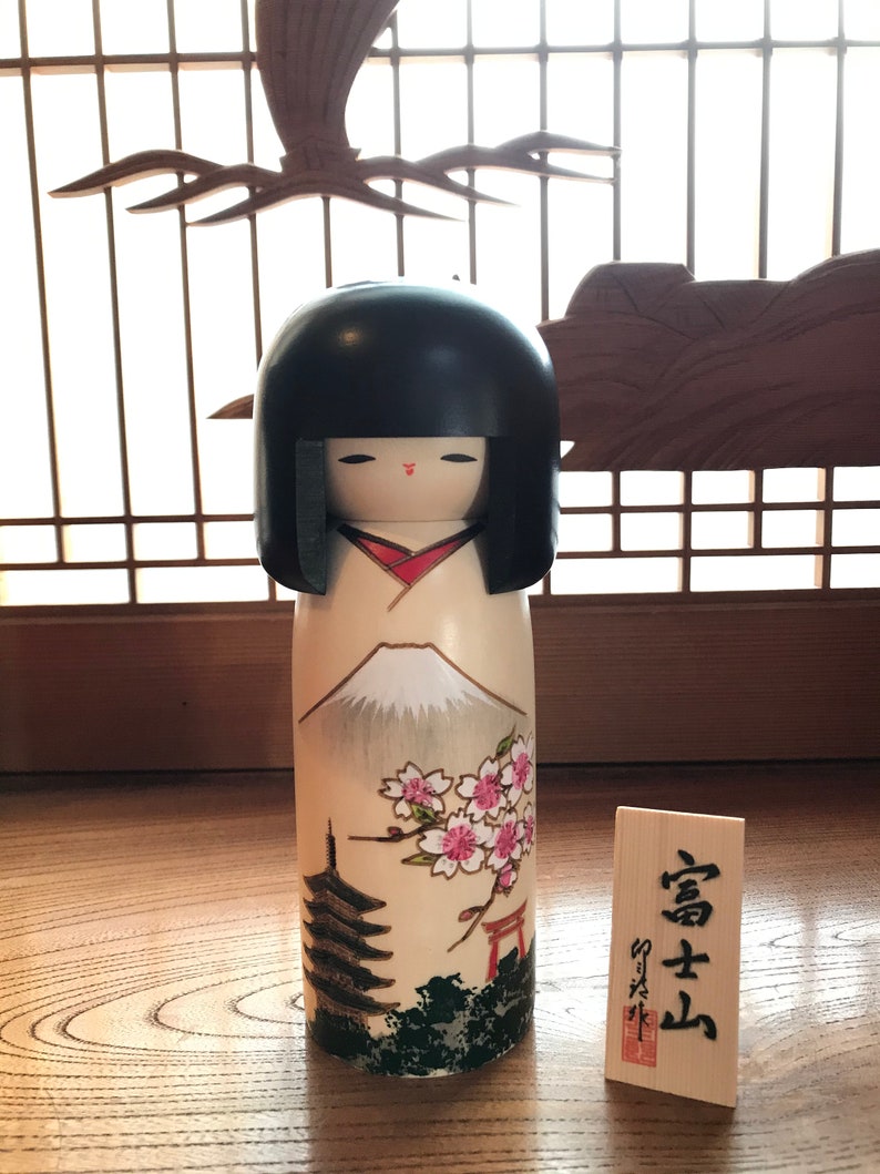 May include: A traditional Japanese kokeshi doll with a black hair, red lips, and a white body with a painted scene of Mount Fuji, cherry blossoms, a pagoda, and a torii gate. The doll is standing on a wooden surface with a small wooden sign that reads "富士山".
