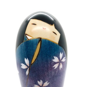 May include: A wooden kokeshi doll with a blue and purple kimono decorated with white flowers. The doll has black hair and a painted face with closed eyes and a smile.
