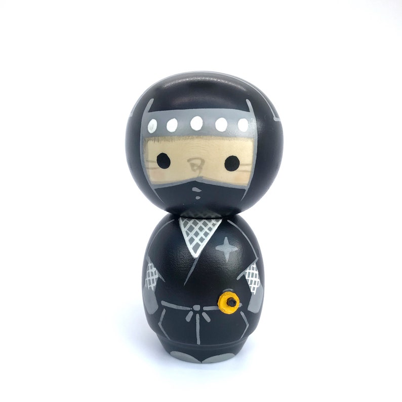 May include: A black and gray wooden doll with a ninja costume. The doll has a black mask, a gray headband with white dots, and a black and gray ninja suit with a star on the chest. The doll is standing on a white background.