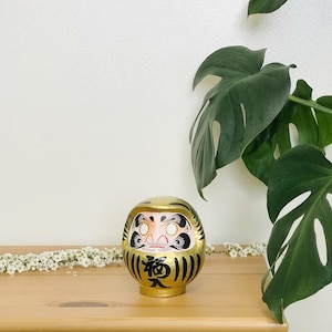 May include: A gold daruma doll with black and white painted details. The doll has a red mouth and white eyes. The doll is sitting on a wooden surface.