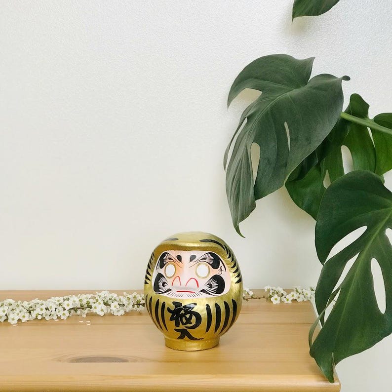 May include: A gold daruma doll with black and white painted details. The doll has a red mouth and white eyes. The doll is sitting on a wooden surface.