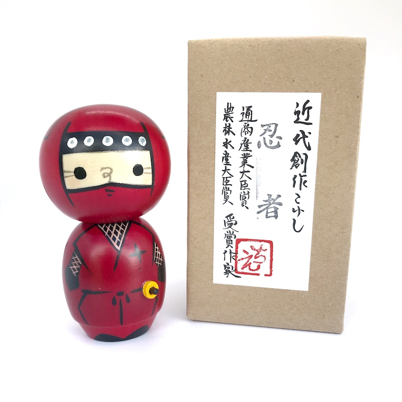 May include: A red and black wooden kokeshi doll, a traditional Japanese doll, depicting a ninja with a white face and black eyes. The doll is wearing a red ninja suit with a black star on the chest and a black belt. The doll is holding a small yellow object in its right hand. The doll is standing on a white surface. The doll is in front of a brown box with Japanese text on a white label.