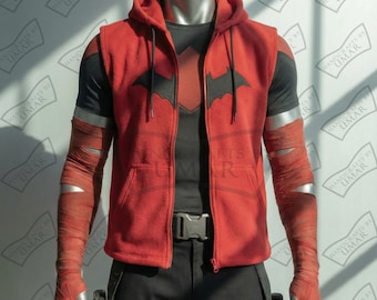 May include: Red hooded vest with a black bat symbol on the chest, worn over a black shirt. The sleeves are wrapped in red bandages with silver accents. The outfit includes black pants and a silver belt buckle. The mannequin is on display.