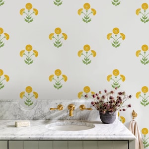 Puede incluir: Un baño con una pared blanca con un patrón repetido de diseños florales amarillos. El tocador tiene una encimera de mármol, un lavabo blanco y accesorios de latón. Un pequeño jarrón de flores secas se encuentra en la encimera.