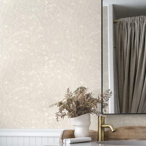 May include: A bathroom scene featuring a cream-colored floral wallpaper. A white vase with dried plants sits on a wooden vanity with a gold faucet. A mirror with a black frame reflects a curtain.