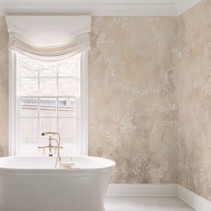 May include: A white freestanding bathtub with gold hardware positioned in front of a window with a white fabric roman shade. The walls feature a beige and cream mural with a painted botanical leaf pattern and white baseboard trim.