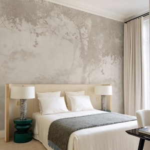 May include: A bedroom with a neutral color palette. The focal point is a bed with white linens, a gray throw blanket, and two white pillows. A tree mural adorns the wall behind the bed. Two lamps with textured bases sit on either side of the bed, and a green side table is on the left.
