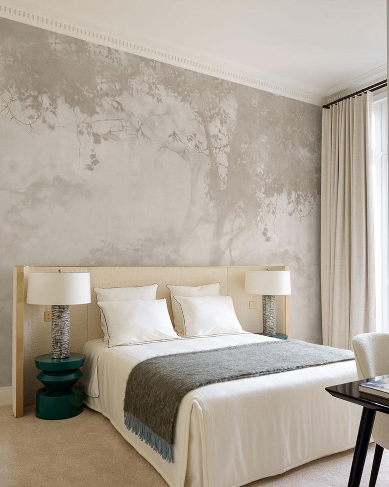May include: A bedroom with a neutral color palette. The focal point is a bed with white linens, a gray throw blanket, and two white pillows. A tree mural adorns the wall behind the bed. Two lamps with textured bases sit on either side of the bed, and a green side table is on the left.