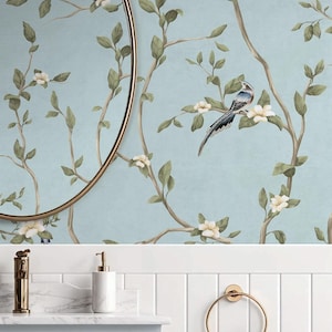 May include: A light blue bathroom with a floral wallpaper featuring a bird perched on a flowering branch. A gold-framed mirror hangs above a white countertop with a gold faucet, soap dispenser, and towel ring. A white towel hangs on the ring.