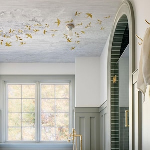 May include: A bathroom interior featuring a ceiling adorned with gold-colored bird decals. The room has a large window, a shower with a glass door, and a gold-colored faucet. The walls are painted in shades of white and gray.